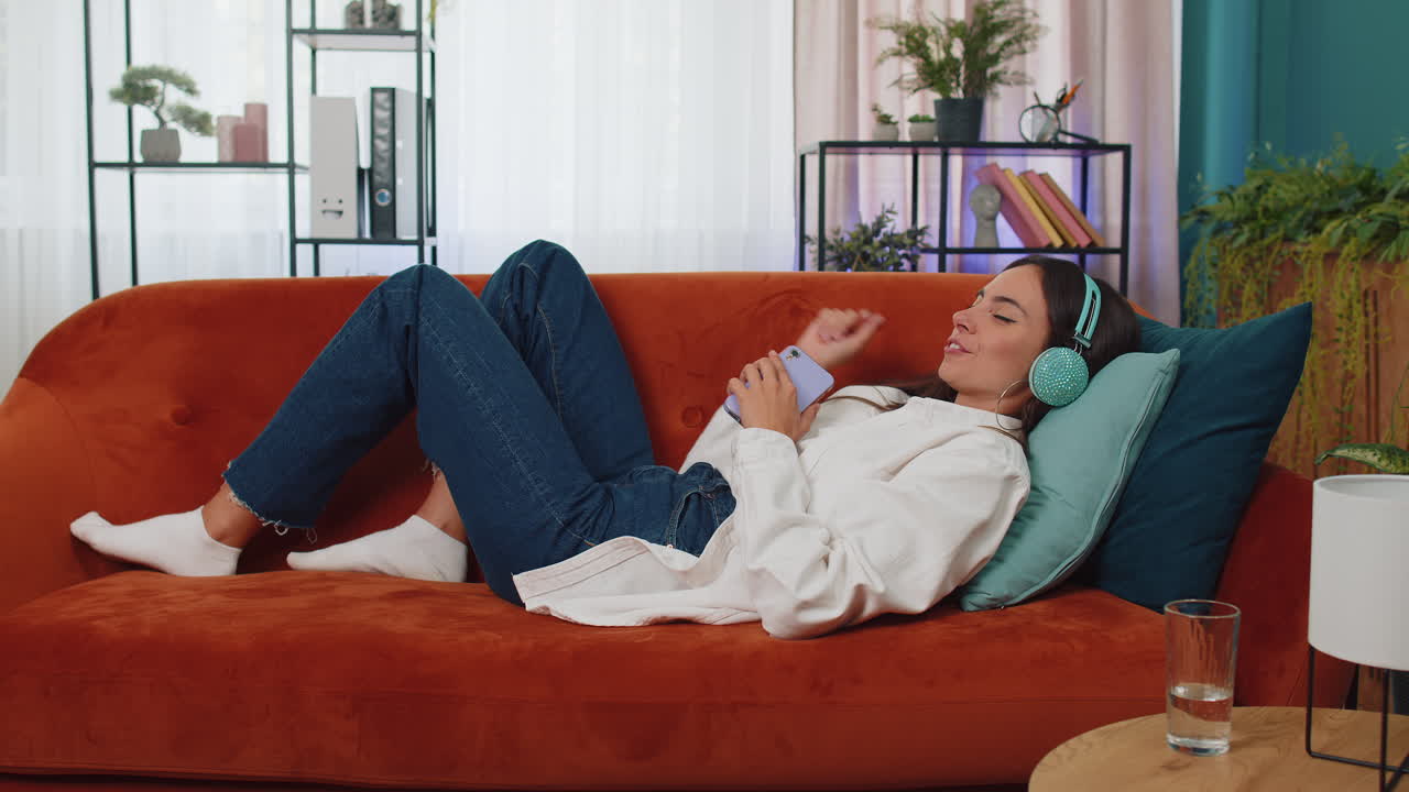 무선 헤드폰을 입은 행복한 젊은 여성이 집에서 소파에 누워서 좋아하는 음악을 듣고 휴식을 취하고 있습니다.