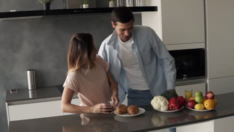 Morning-at-the-kitchen-counter,-fast-breakfast-before-work---croissants,-juice-and-coffee-for-breakfast.-Handsome-husband-came-to-the-kitchen,-welcomes-his-wife-with-a-kiss,-sipping-an-orange-juice,-wife-is-drinking-coffee-or-tea-from-a-big-mug