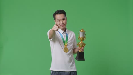 hombre asiático con una medalla de oro y un trofeo señalando y mostrando los pulgares hacia abajo gesto para ridiculizar al perdedor en el fondo de la pantalla verde en el estudio