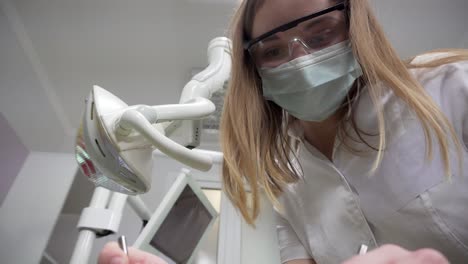 Junge-Zahnärztin-Mit-Maske-Untersucht-Den-Patienten-Mit-Werkzeugen,-Steht-Auf-Einem-Patienten,-Blickt-In-Die-Kamera-Und-Sieht-Das-Gesicht-Des-Zahnarztes