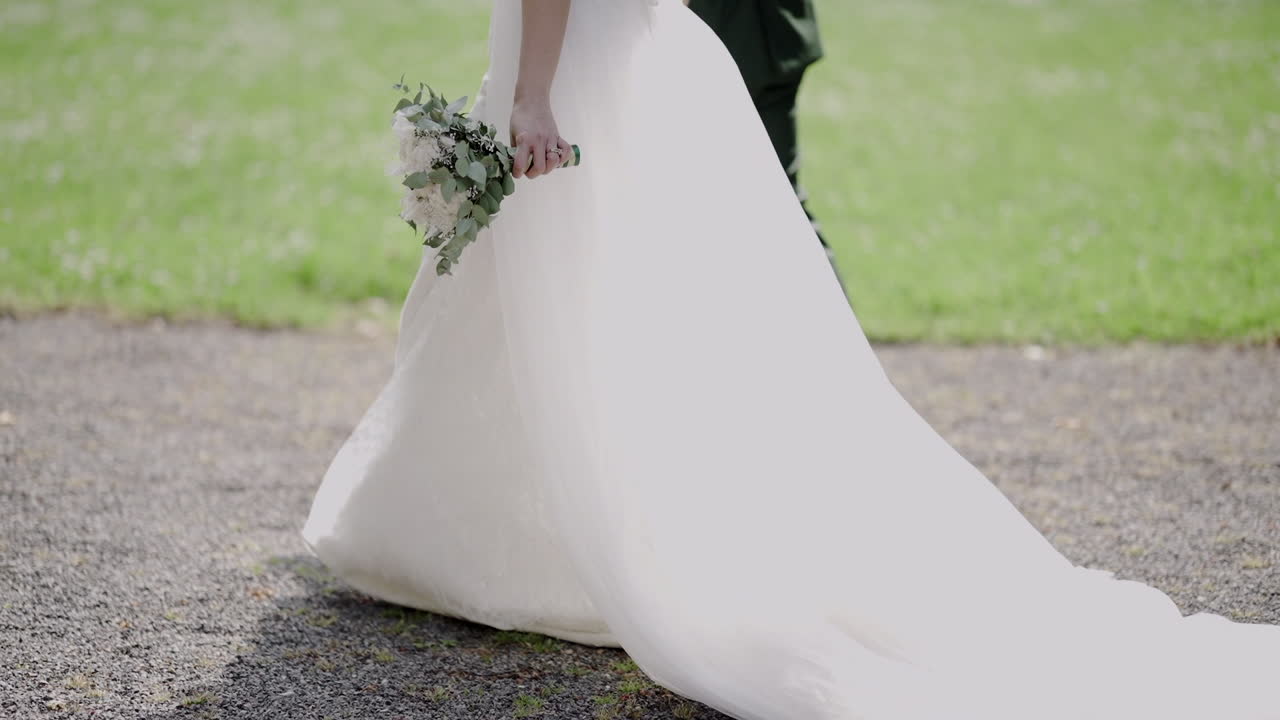 Brud med buket går med gom på en sti, hendes hvide kjole slæber elegant hen ad jorden.