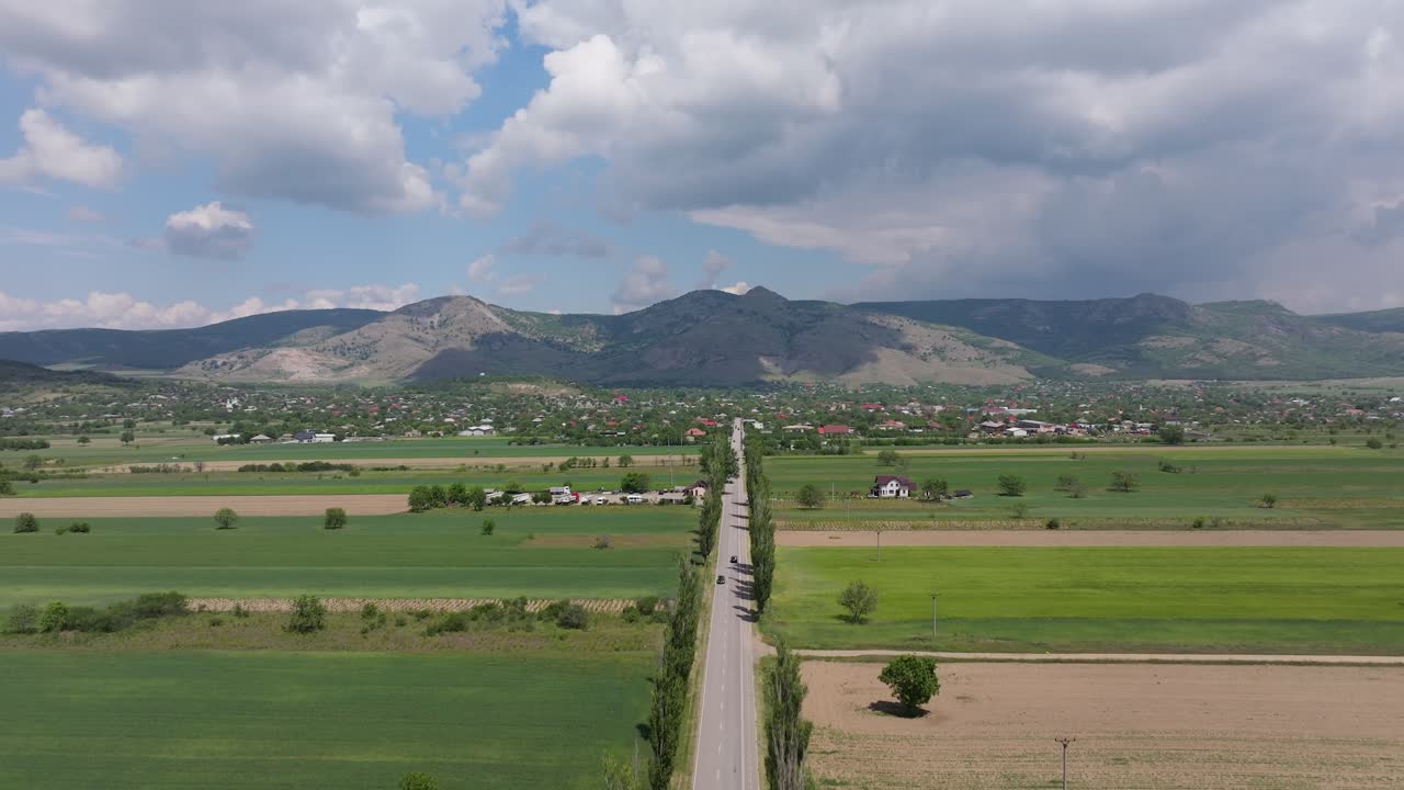 Rising drone shot gradually revealing a straight country road stretching into the distance toward a backdrop of rolling mountains. Leading line through latge green fields