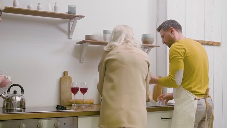 Rear View Of A Man Washing The Family Dinner Dishes At The Sink In The Kitchen While A Mature Women Helping Him