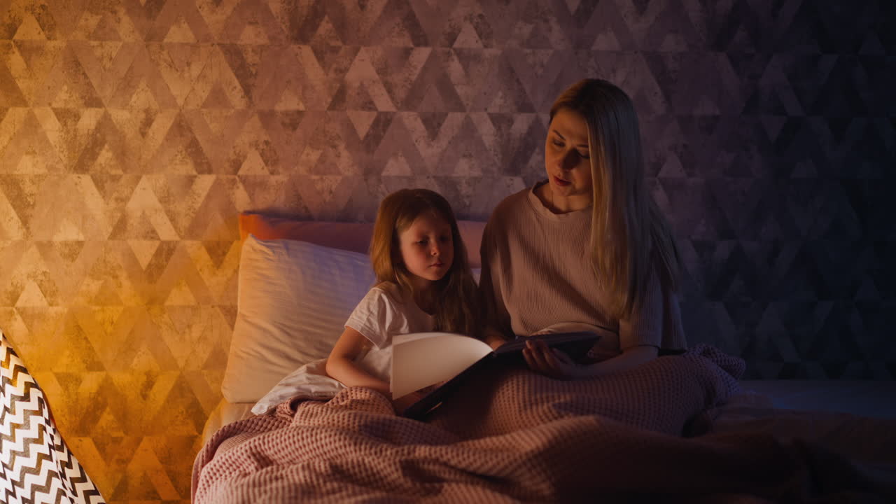 Mother reads fairy tale to daughter sitting together on bed