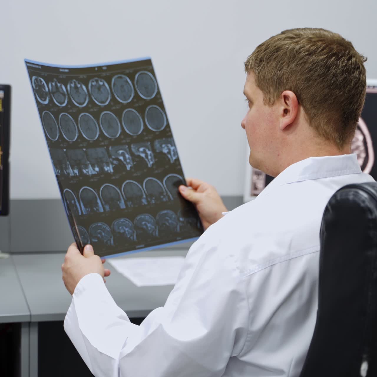 Doctor in white robe sitting his back to the camera holding the MRI scan of brain. Expert consulting to a colleague showing him a shot
