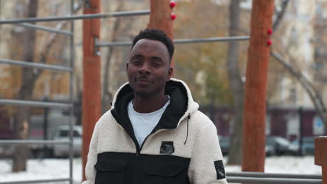 young adult smiles warmly with head slightly tilted as cold breath is visible in crisp winter air, wooden structure and blurred buildings in background