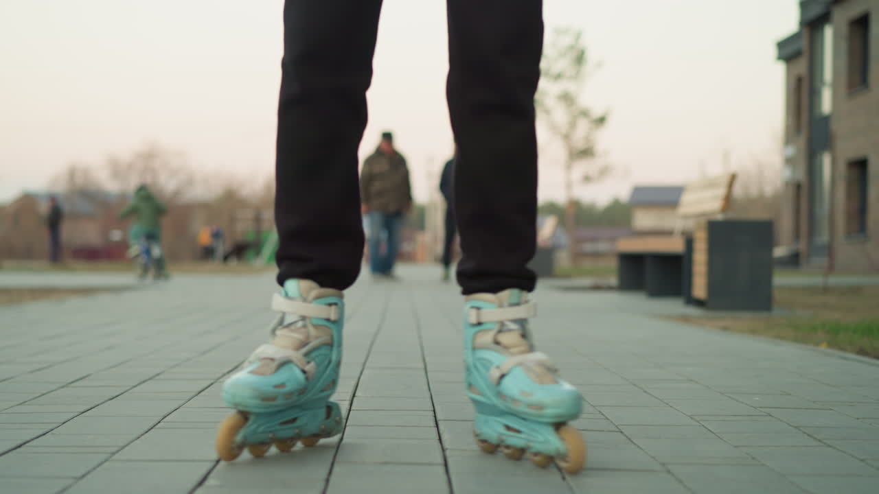 A woman is rollerblading along a paved path in a park, wearing a peach jacket and black trousers. The background reveals a lively scene with other park-goers, including a child on a bicycle and people