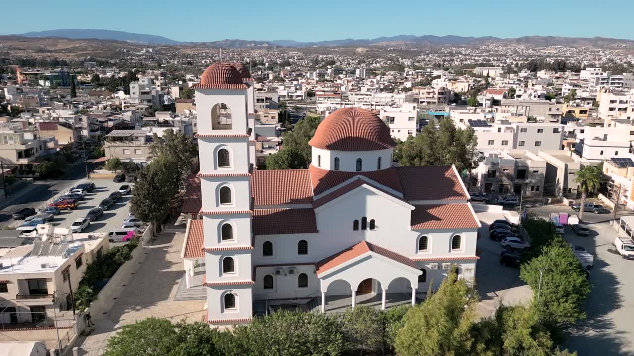 Una chiesa si erge alta in un villaggio. Le case circondano la chiesa e le montagne sono visibili sullo sfondo. La luce intensa del giorno mostra i dettagli dell'edificio e dell'area.
