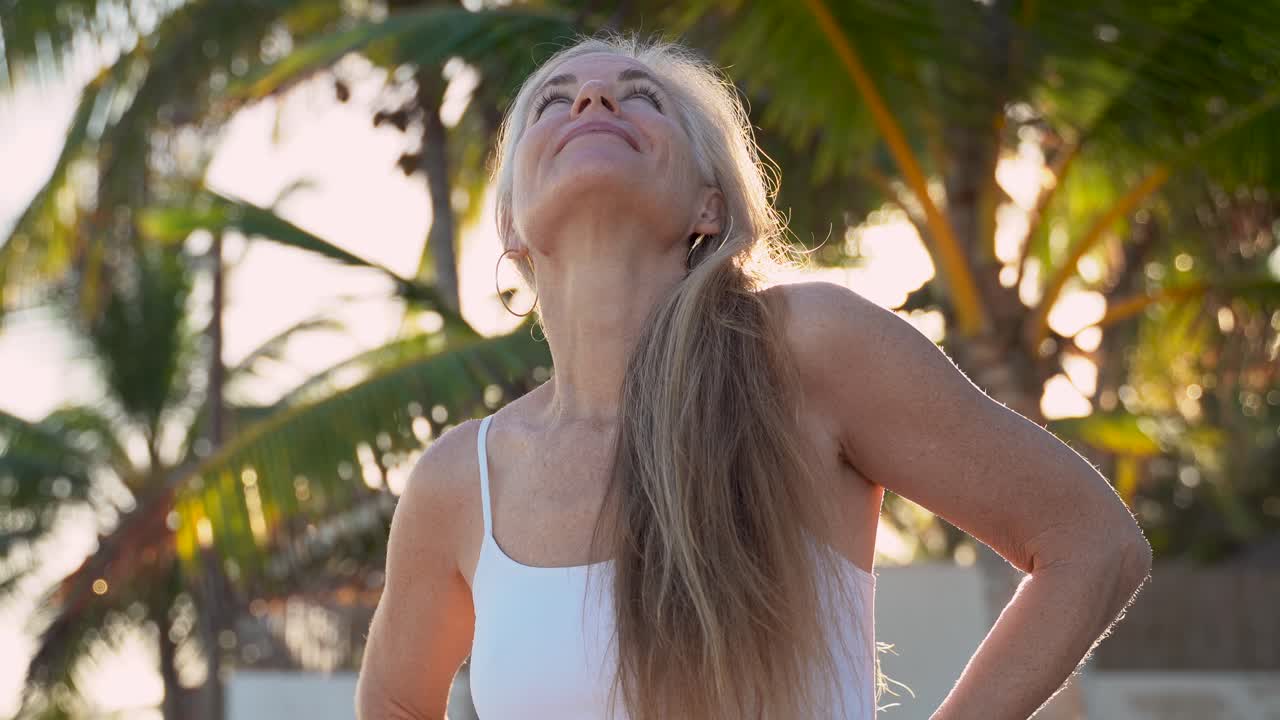 Bella donna matura guarda la telecamera mentre il sole scintilla dietro di lei tra le palme tropicali