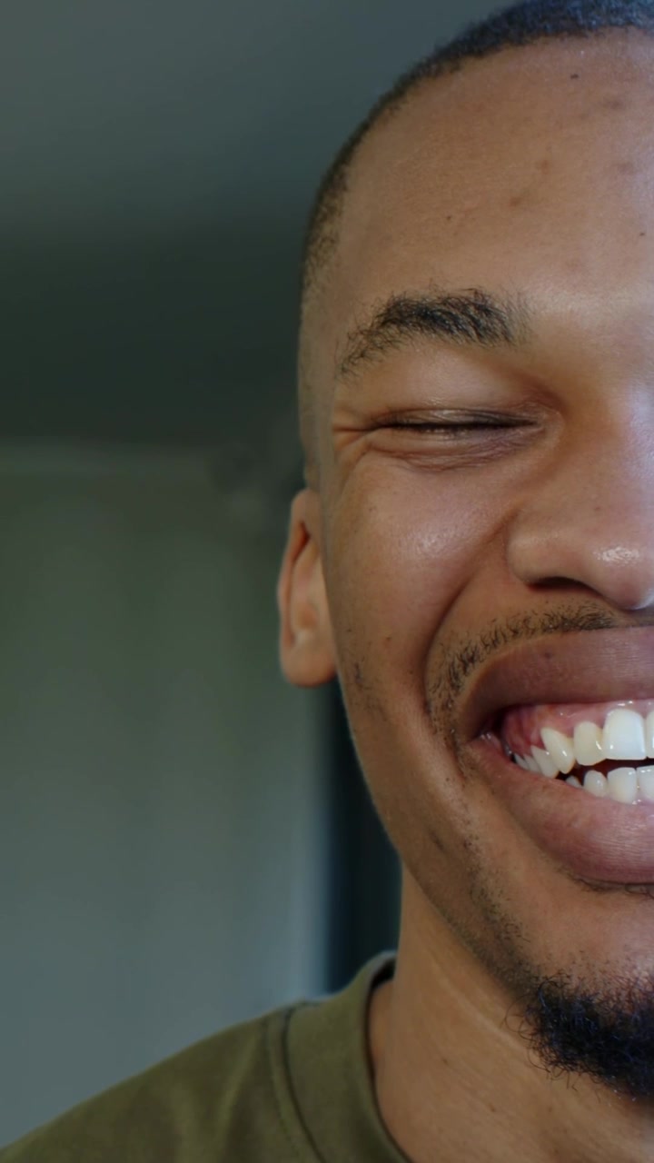Vertical video: Smiling man moving into broad laugh when camera records at home, in olive tee