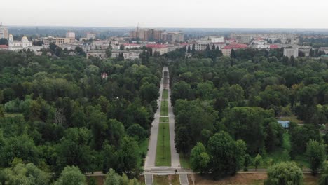 Aerial-View-of-Sidewalk-Going-Through-Park-Surrounded-by-Trees-Leading-to-City-in-Background