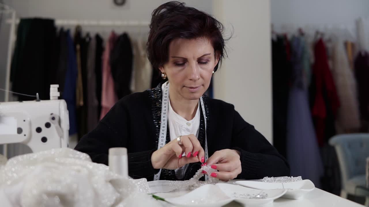 Portrait of a seamstress working on details with wedding dress. Holding needle and thread and decorating exclusive wedding dress with decorative stones. Sewing elegant wedding dress. Hand made. Tailor's studio