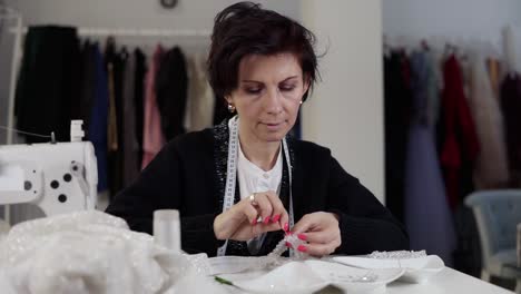 portrait of a seamstress working on details with wedding dress. holding needle and thread and decorating exclusive wedding dress with decorative stones. sewing elegant wedding dress. hand made. tailor's studio