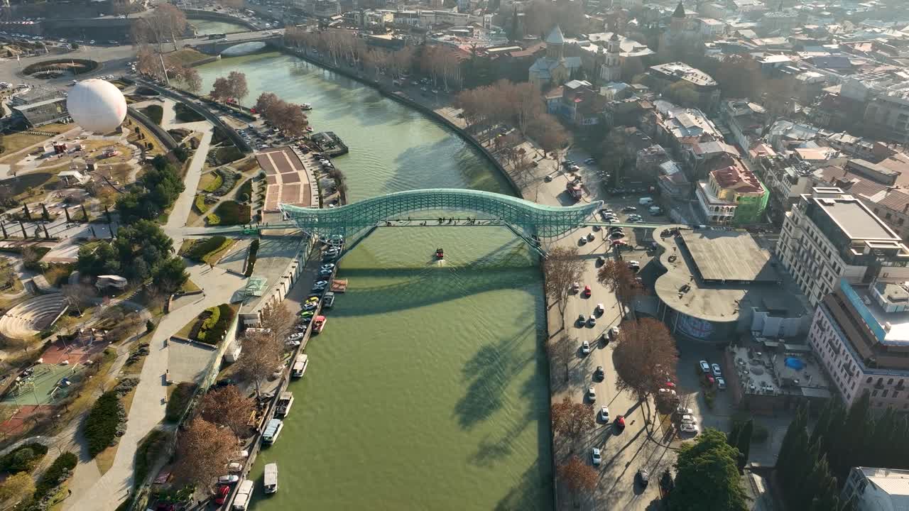 close aerial shot of the Bridge of Peace, a modern glass pedestrian bridge over the Kura River in Tbilisi. The photo captures the reflection of sunlight on the green water