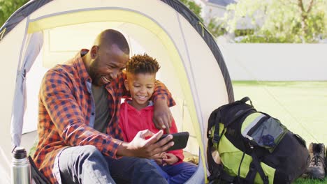 Happy-african-american-father-and-his-son-sitting-in-tent-and-using-smartphone-in-garden