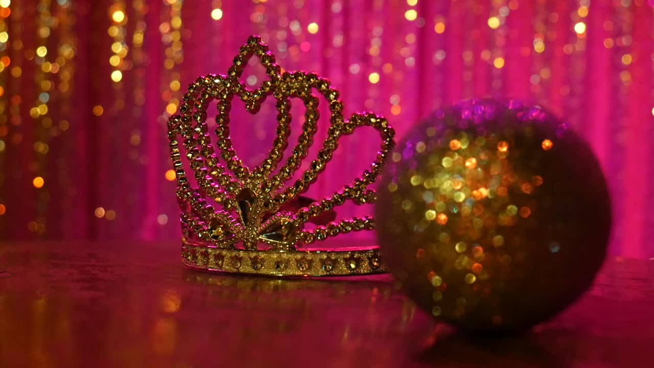 A gold sparkling tiara sits in front of a pink background