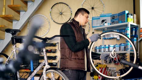 self-employed mechanic is repairing bicycle wheel with wrenches professional instruments while working in small workshop. young man is listening to music with earphones.