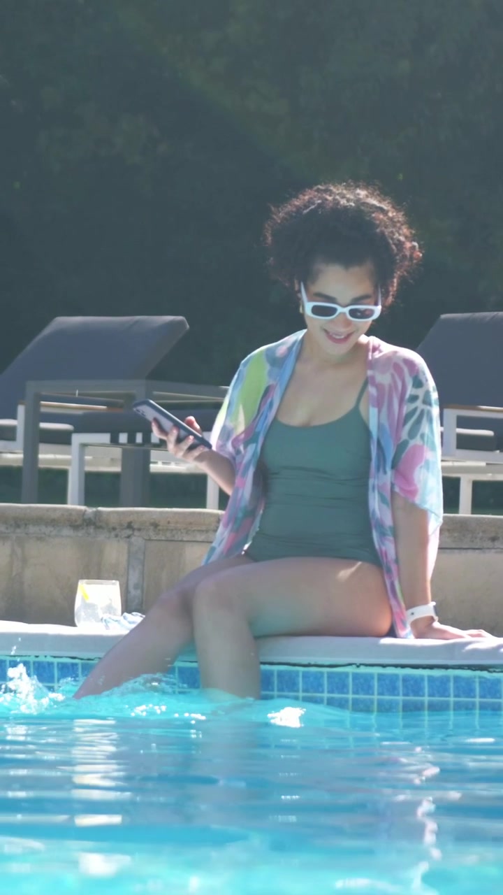 Vertical video: Checking phone, woman in green swimsuit dipping legs in pool at resort, cooling off