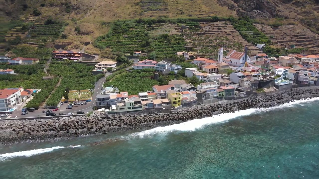 Ripresa con drone che cattura la stretta striscia di terra dove si trova il villaggio di Paul do Mar tra scogliera e mare. La geografia unica evidenzia l'ingegno dell'insediamento in un terreno difficile.