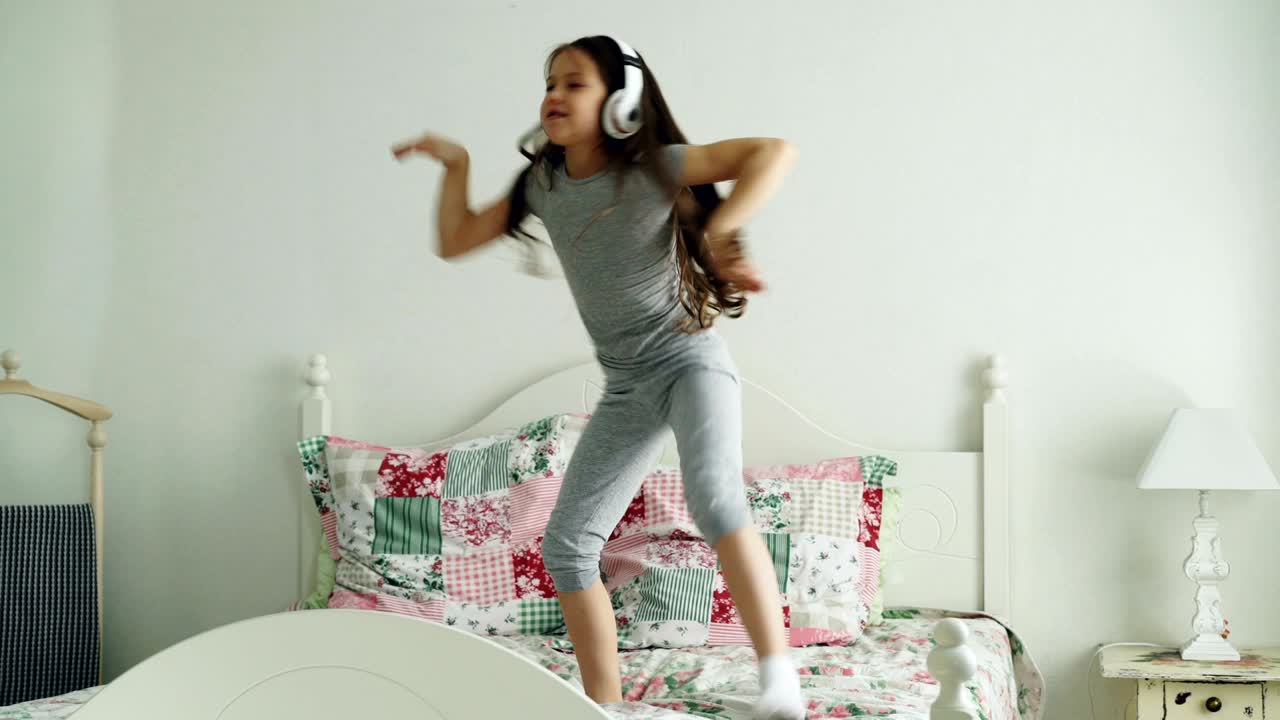 Simpatica e adorabile bambina con cuffie wireless che balla e si diverte sul letto di casa, in una camera da letto accogliente.
