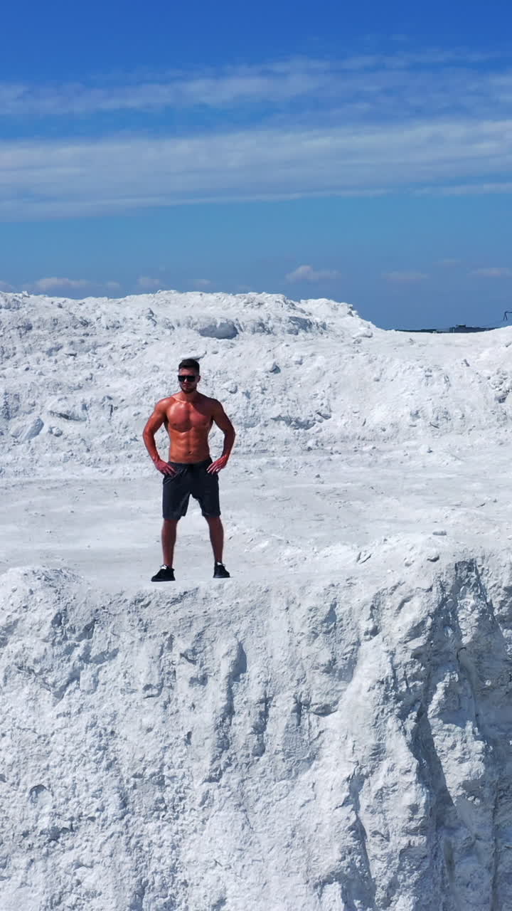 Drone view of a strong man in shorts without shirt on white hill. Young muscular man in good physical shape wearing sunglasses standing in nature. Camera moves up. Vertical video