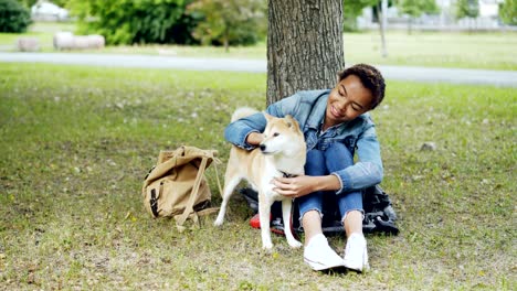 young african american woman is caressing adorable puppy resting in the park on green lawn and talking to pet with kindness and adoration. people and animals concept.