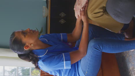 Vertical-Video-Of-Female-Care-Worker-In-Uniform-Holding-Hands-Of-Senior-Man-Sitting-In-Care-Home