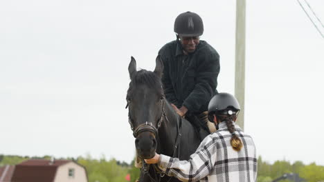 feliz joven montando, sonriendo y montando un caballo negro con la ayuda de una joven