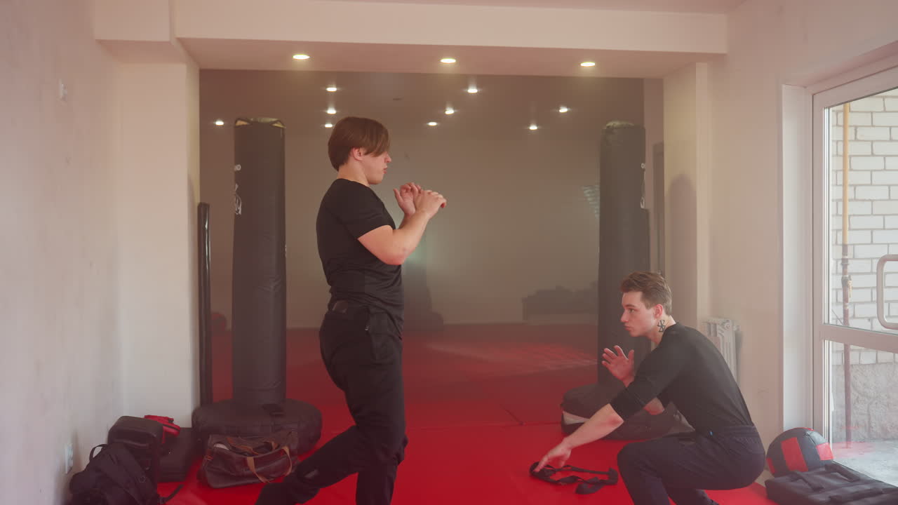 Karate practitioners facing each other in gym during martial arts training session, one in fighting stance with raised hands preparing for sparring while other observes calmly, dodging