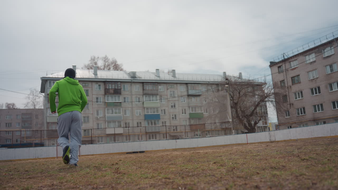 mentor canoso de edad avanzada gesticulando y aconsejando mientras un jugador joven corre por el campo, chaqueta desgastada y postura tranquila, entorno de patio urbano, consejo cálido y presencia constante en el aire fresco