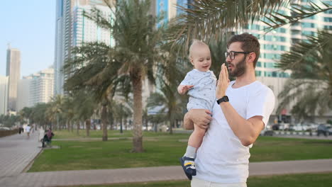 Vater-Spielt-Mit-Söhnen-Im-Sommer-In-Einer-Modernen-Stadt,-Hält-Ihn-Und-Umarmt-Ihn,-Stehend-In-Einem-Weißen-T-Shirt-Und-Shorts