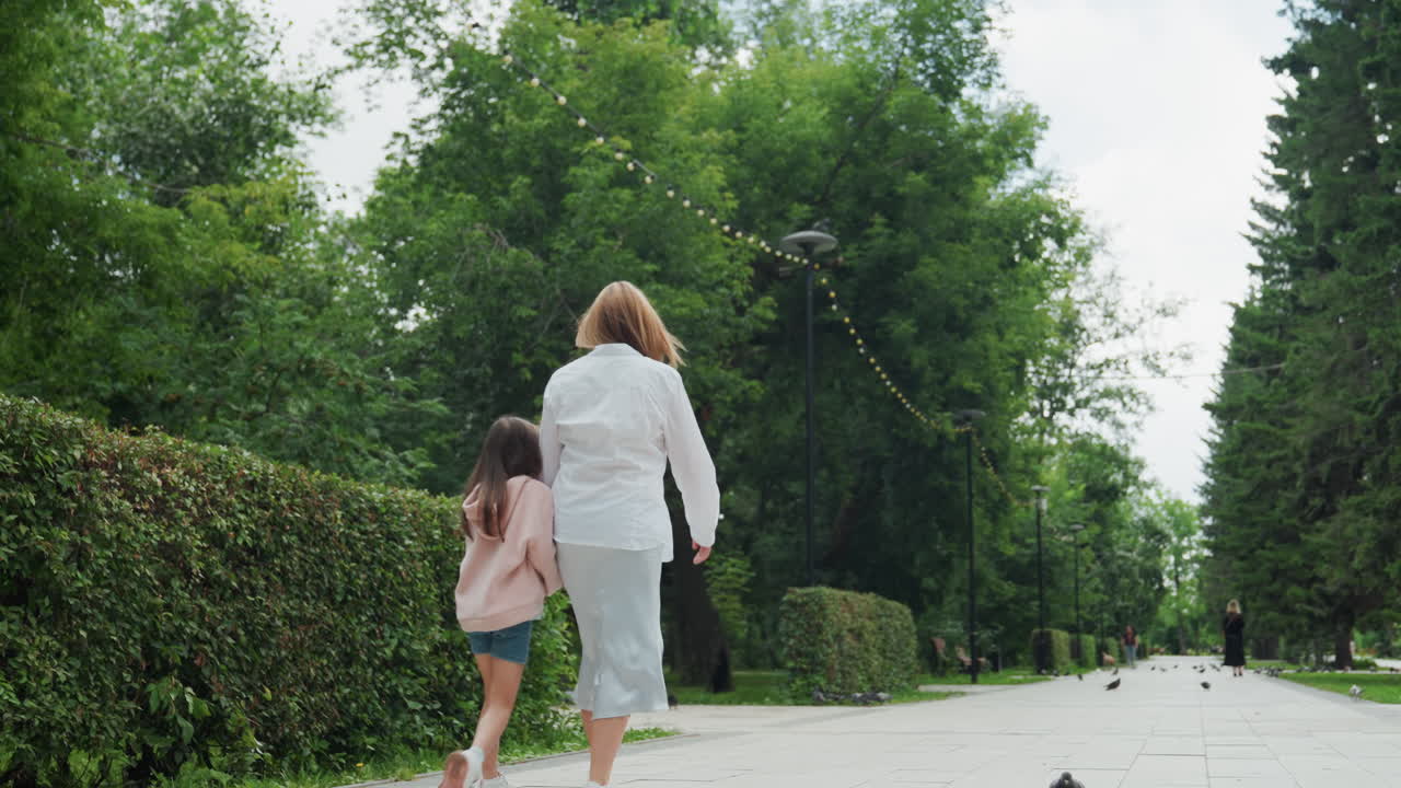 Kaukasische moeder en dochter lopen weg onder hoge bomen, achteraanzicht van moeder die kind leidt over een met bomen omzoomde laan, contemplatieve stemming, zomerse natuur, lange perspectief reis en verbinding.