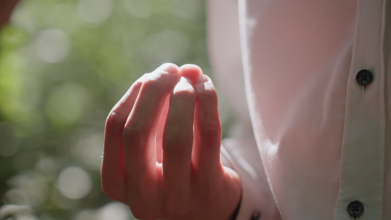 Close up of man hand in white shirt with blurred green forest background, fingers slightly curled, natural sunlight highlighting skin texture creating atmospheric view of human detail
