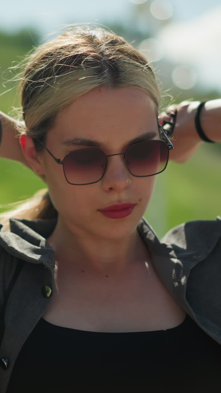 회색 재 에 검은 을 입은 여성, 선글라스를 입고, 큰 헤어 클립으로 머리카락을 자르려고 노력하고, 그녀의 초점은 그녀가 은 날에 서있는 동안 그녀의 머리카락의 배열에 있습니다, 언덕의 흐릿한 시야와 함께