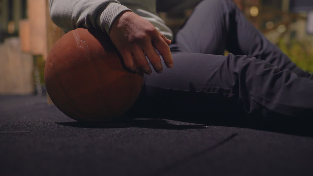 Natlig basketballspiller hviler med basketball, urban tagterrassebane oplyst af varme bylys, mandlig atlet iført hættetrøje og bukser sidder på asfalt, hånd griber bolden, sneaker-sål synlig, eftertænksom