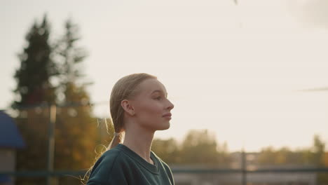 side view of lady in green sweater playing volleyball outdoors, adjusting her stance to serve, with a clear volleyball net in the foreground and a background of greenery and buildings