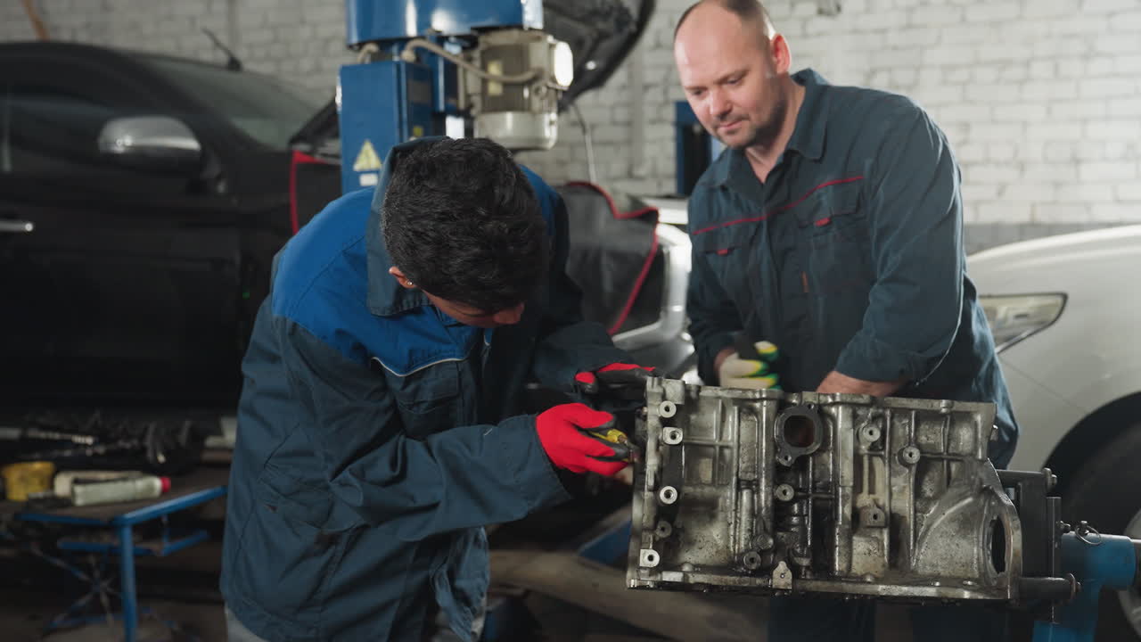 dos mecánicos en uniformes azules trabajando en un motor en un taller de automóviles, uno raspando el motor mientras el otro suelta los pernos, rodeados de herramientas y piezas de automóviles