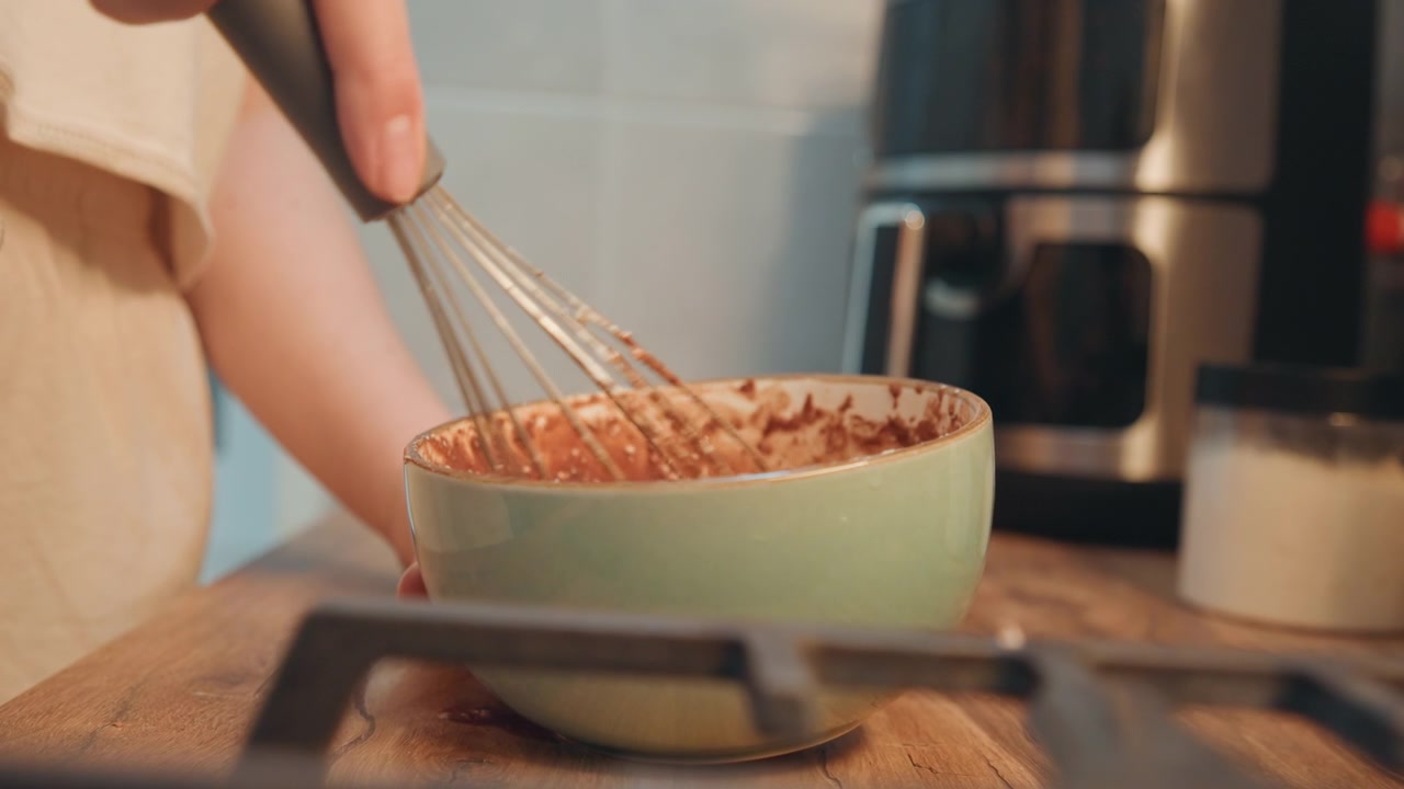 Ampia inquadratura di impasto al cioccolato mescolato, chef casalinga con grembiule al bancone vicino alla macchina del caffè, luce del mattino, routine di colazione informale, consistenza liscia dell'impasto, scena di cucina che evoca uno stile di vita rilassato.