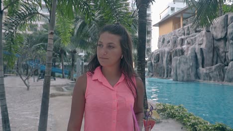 girl with drink in hand walks near palms and swimming pool