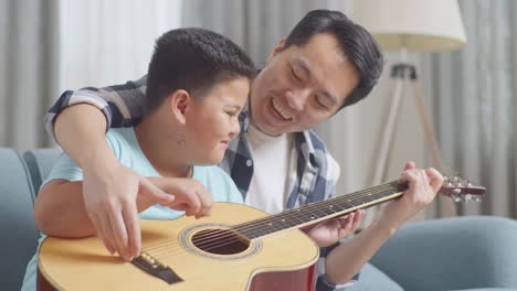 close up of asian father teaching his son singing and playing the guitar on sofa at home