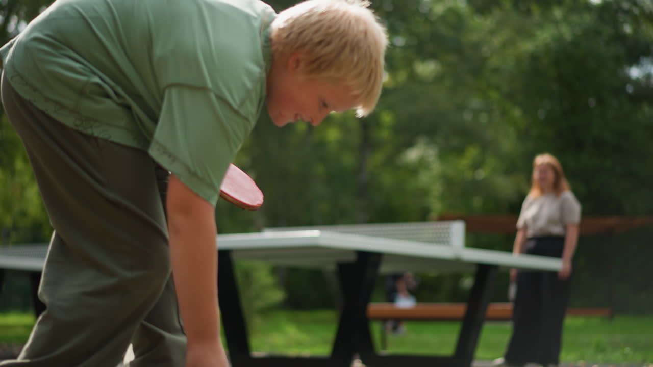 Afslappet spil mellem ung dreng og kvinde, der smiler og har en sjov udveksling på banen med en afslappet og venlig atmosfære, muntert spil mellem ung mand og kvinde med tilskuere og bænke i nærheden