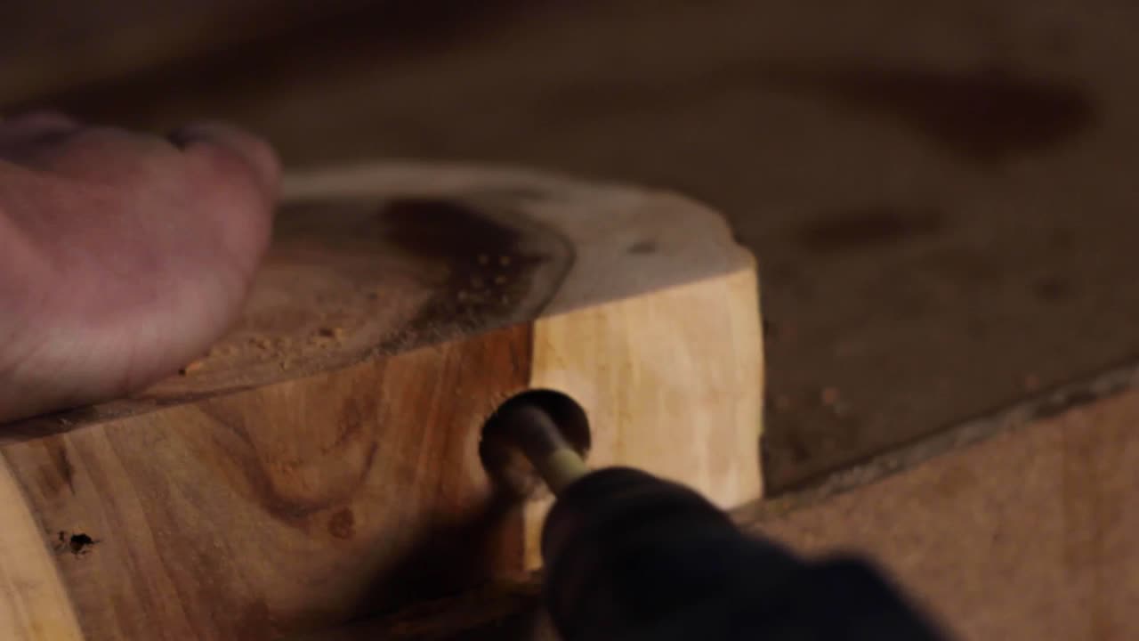 Close up of a carpenter drilling a circular hole into a large slab of hardwood, held down by the carpenter's bare hand