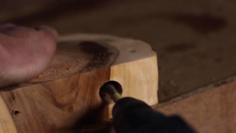 Close up of a carpenter drilling a circular hole into a large slab of hardwood, held down by the carpenter's bare hand