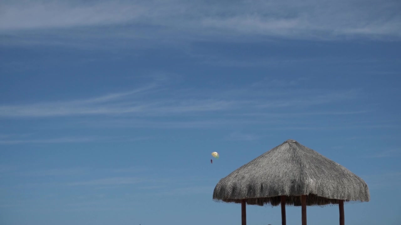 Rifugio da spiaggia con tetto di paglia sotto un cielo azzurro e aperto, con un paracadute ascensionale in lontananza sopra una costa tropicale e un'atmosfera marittima tranquilla.