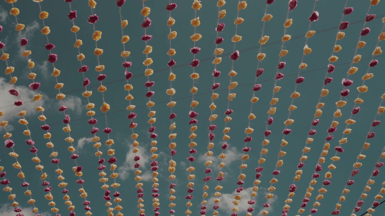 Strings of handmade paper flower garlands in orange and maroon hang against blue sky at arraial festival