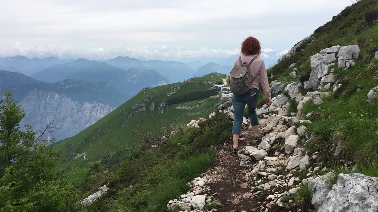 una toma siguiendo a una mujer que camina por un sendero estrecho en la cima de una montaña, con un fondo de montañas y nubes bajas
