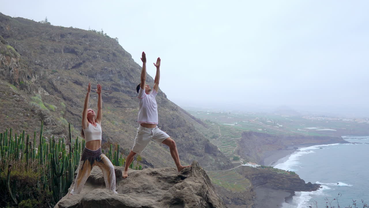 Balanced at a cliff's edge, a man and woman raise their hands, inhaling ocean air during a yoga session while gazing at the sea