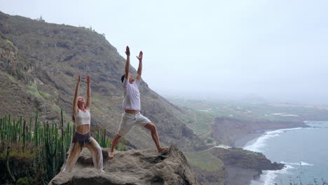 equilibrados en el borde de un acantilado, un hombre y una mujer levantan las manos, inhalando el aire del océano durante una sesión de yoga mientras miran el mar