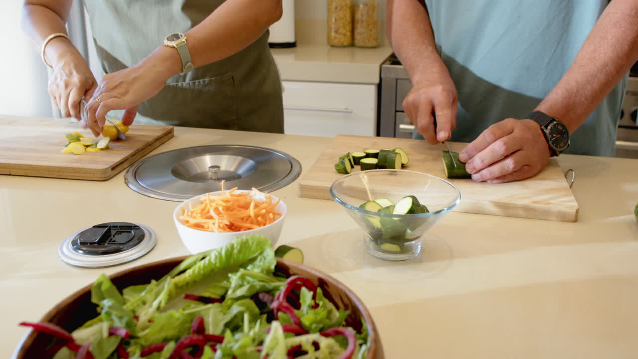 Senior diverse couple preparing fresh salad together in modern kitchen, enjoying cooking, at home