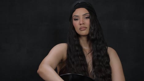 A Model with Black Wavy Hair and Black Clothes Sits on a Chair and Tilts Her Head to One Side. A model with black wavy hair and black clothes sits on a chair and tilts her head to one side del with black wavy hair and black clothes sits on a chair and tilts her head to one side. High quality footage